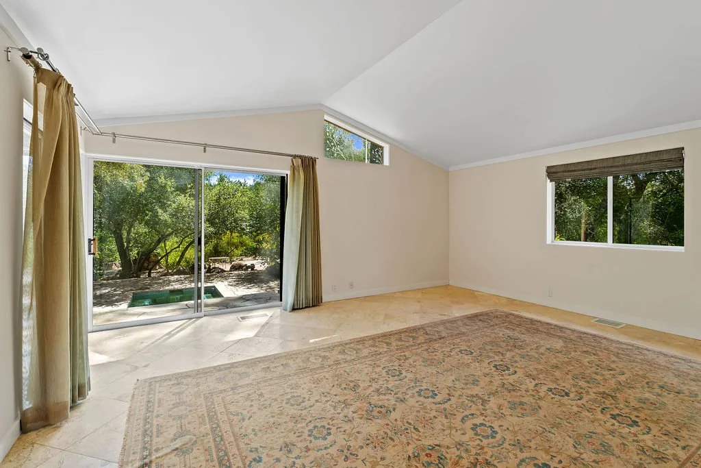 Empty living room with large glass sliding doors, a window with blinds, and a patterned rug, overlooking a backyard with trees.