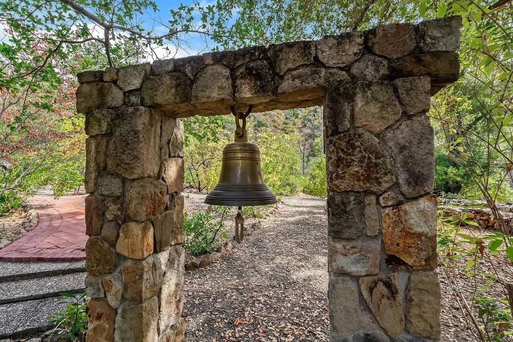 A stone archway with a hanging bell in a garden setting, surrounded by trees and greenery.