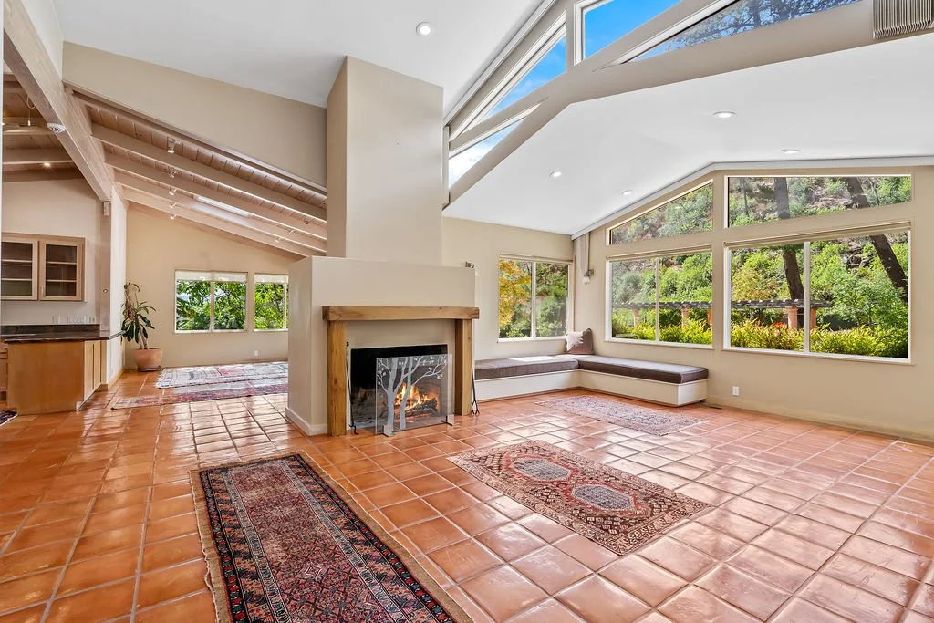 Bright living room with sloped ceiling, large windows, tiled floor, and a fireplace with a wooden mantel.