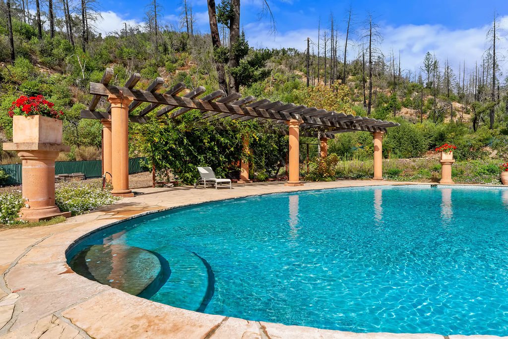 An outdoor swimming pool with clear blue water, surrounded by a stone deck, in a garden with potted red flowers, green trees, and a hillside with dry trees under a partly cloudy sky.