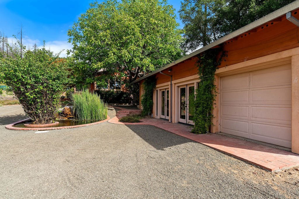 Front view of a residential house with a garage, surrounded by lush trees and a landscaped yard with bushes and plants.