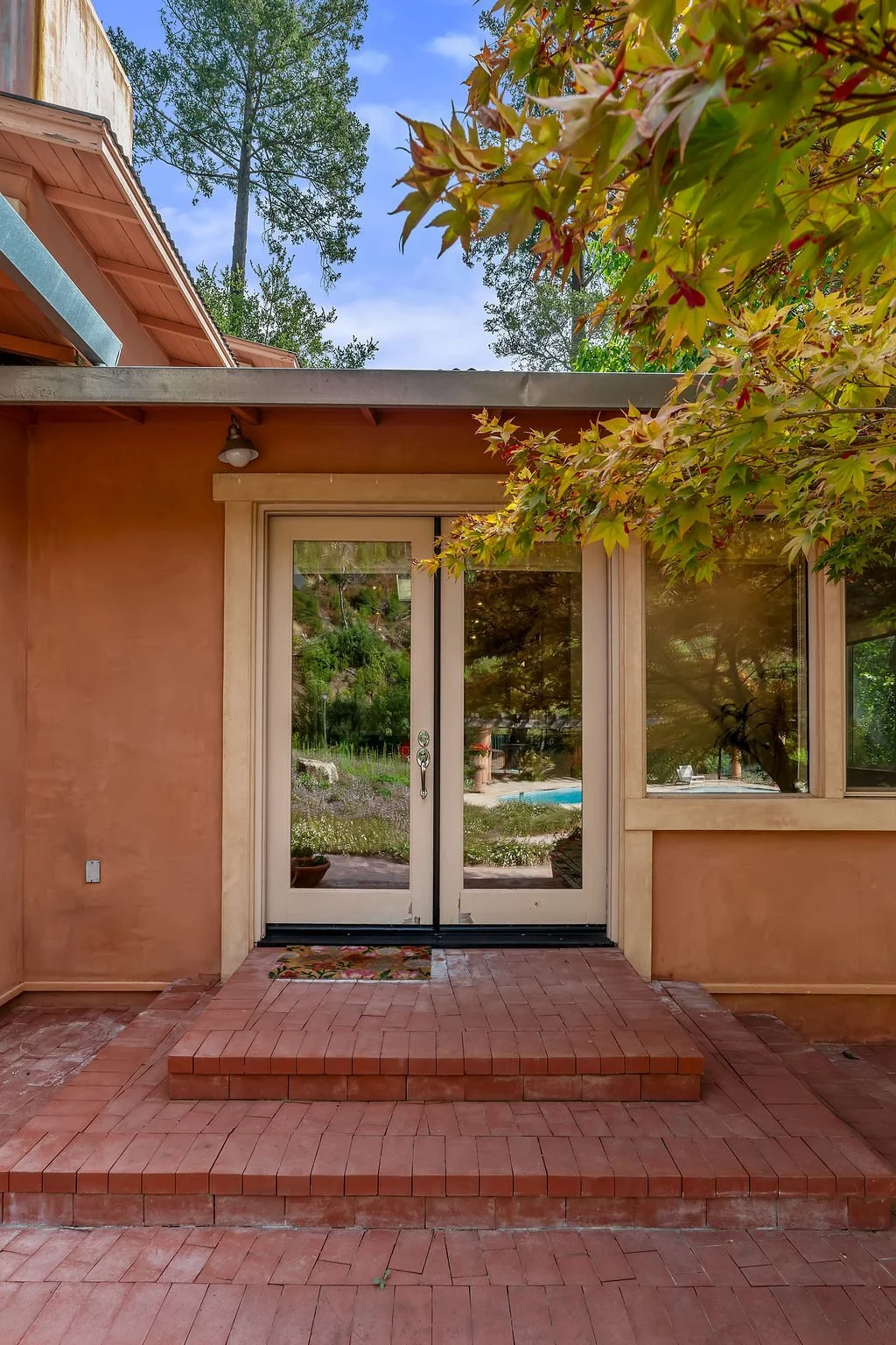 The image shows a back patio with brick steps leading to glass double doors. There is a tree with green leaves and a branch with reddish leaves partially covering the top right corner. The background includes a view of a backyard with a swimming pool