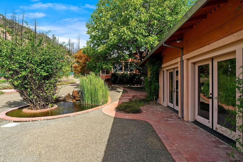 Outdoor scene with a brick pathway, lush green trees, and a building with glass doors and a wooden exterior.