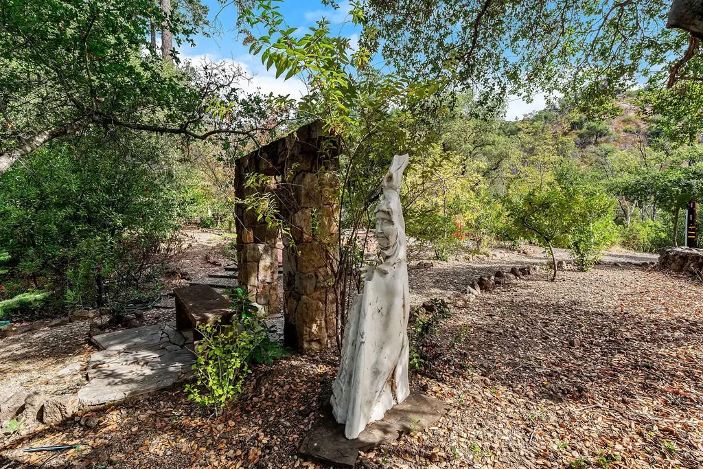 A white sculpture of a person with facial features and a raised arm, located in an outdoor garden area with trees, shrubs, and gravel ground.