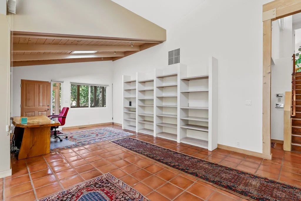 Living room with terracotta tile flooring, white walls, an open staircase, and a wooden ceiling. Features include a red office chair at a wooden desk, or rugs, and white built-in bookshelves against the wall. Large window with outdoor greenery visibl