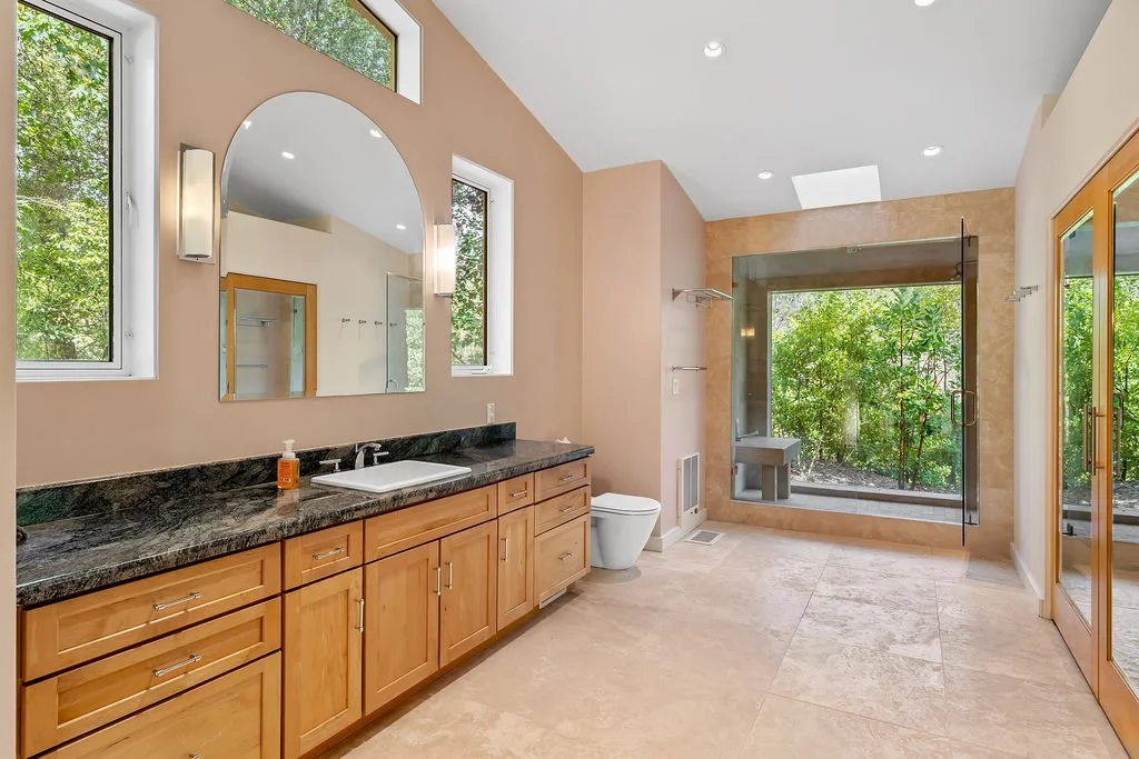 A spacious bathroom with a vanity featuring a dark granite countertop, wooden cabinets, and a large mirror. There are three windows providing natural light, a modern white toilet, and a walk-in shower with glass doors and a built-in seat, all with a 
