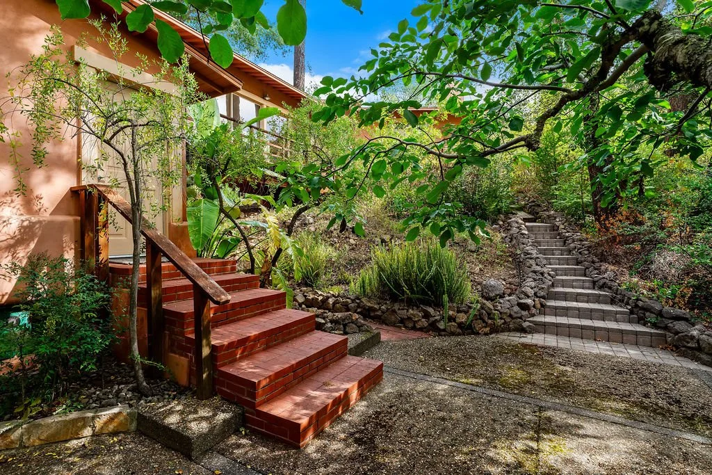 Brick stairs leading to a house entrance with surrounding lush green trees and plants, an ascending stone walkway on the right side, and a clear blue sky overhead.