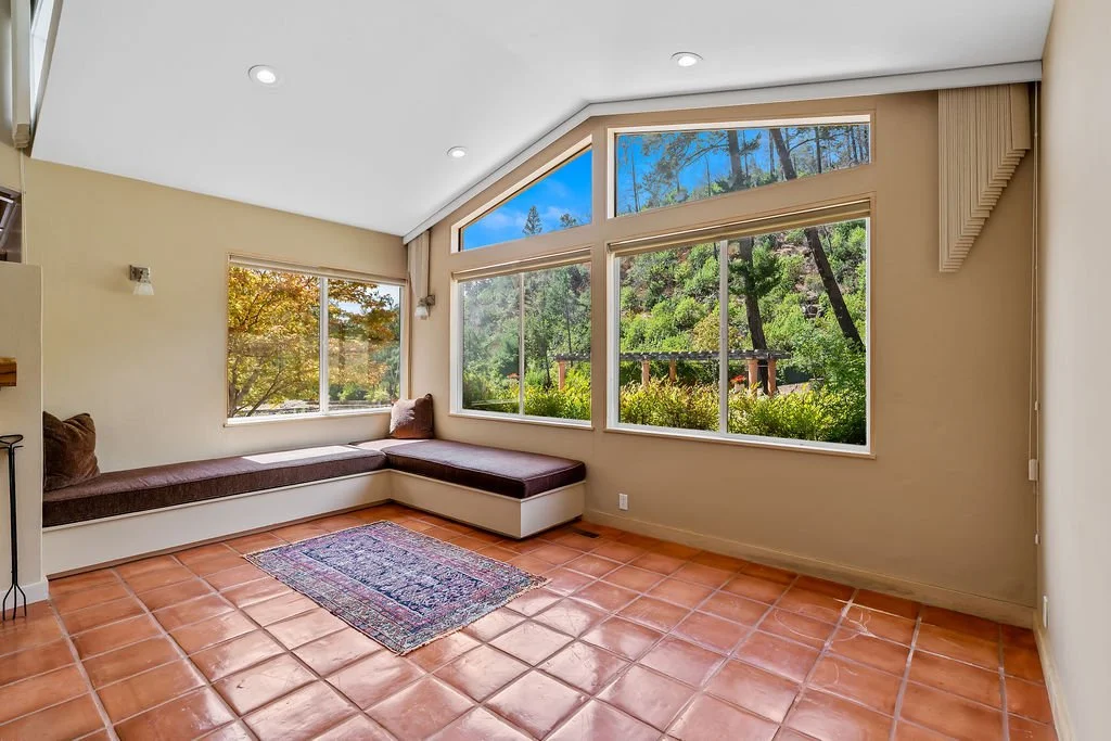 Living room with large windows showing trees and greenery outside, built-in bench with cushions, terracotta tile floor, a small rug, and beige walls.