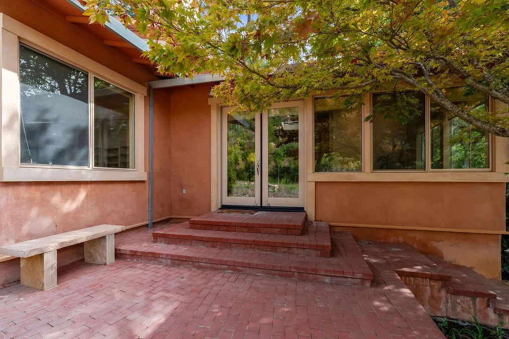 A house entryway with a brick patio, stairs, and glass door, shaded by a large leafy tree.