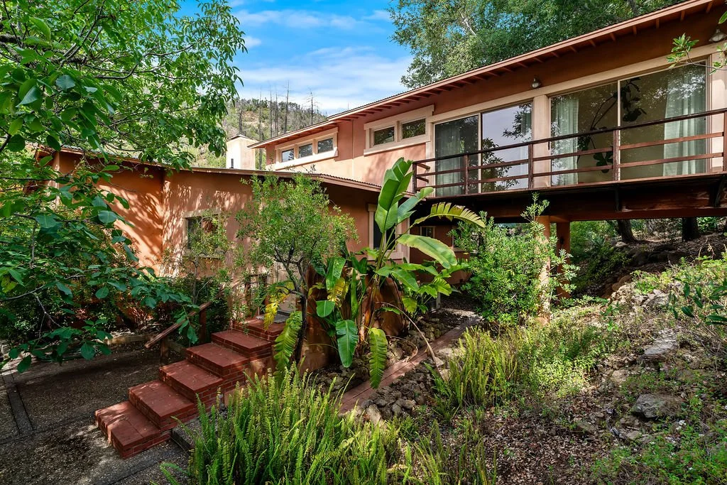 A mid-century modern house with large glass windows and a wooden balcony, surrounded by lush green trees and plants on a hillside.
