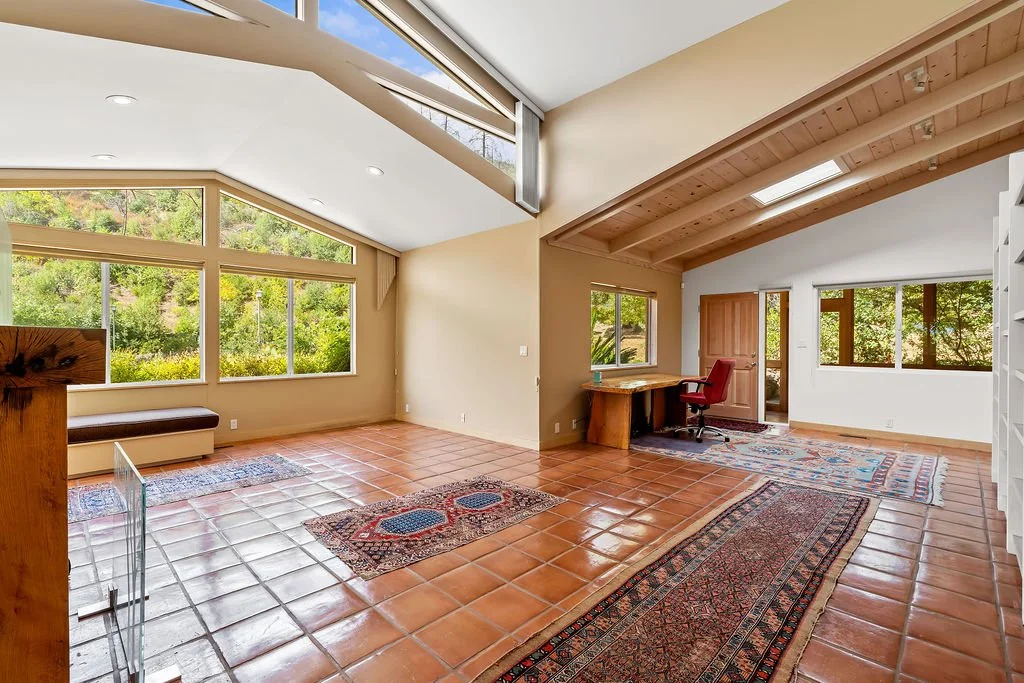 Spacious living room with large windows, terracotta tiled floors, and a wooden ceiling. Contains rugs, a built-in bench, and a corner desk with a red chair.
