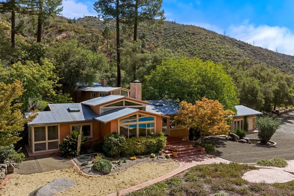 A single-story house with a modern design, metal roof, and large windows, surrounded by lush trees and shrubbery, situated in a hillside landscape with mountains in the background.