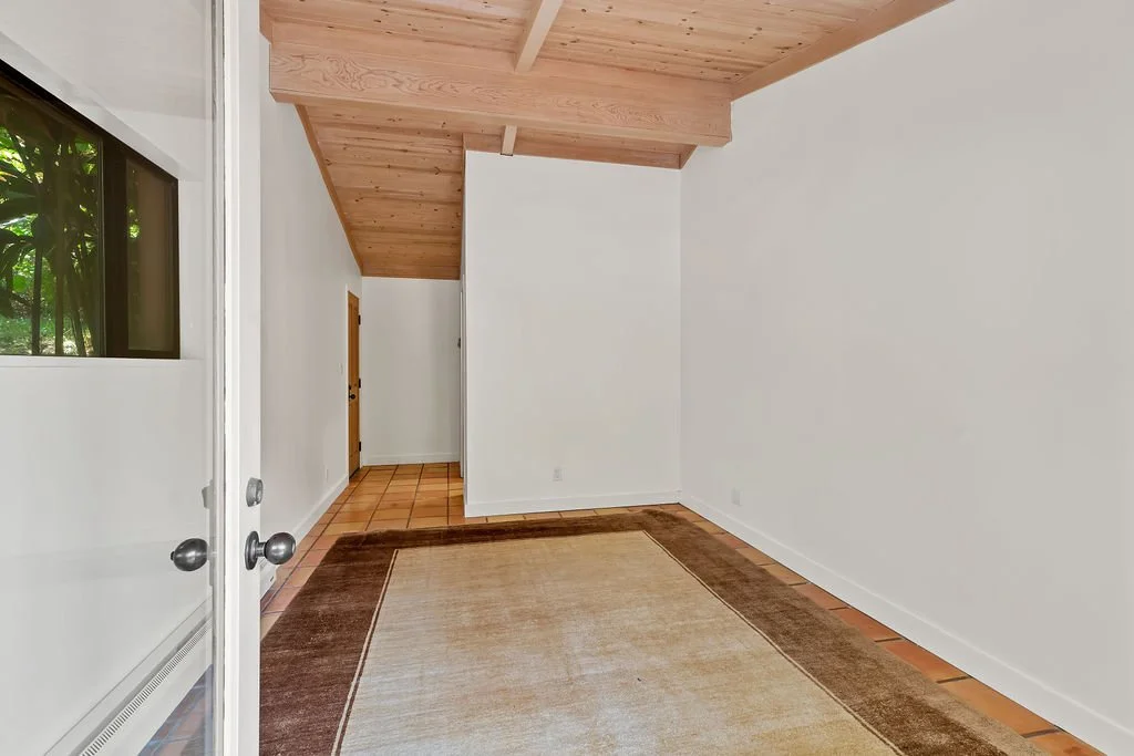 Empty room with white walls, wooden ceiling, tiled and hardwood flooring, and a large window with greenery outside.
