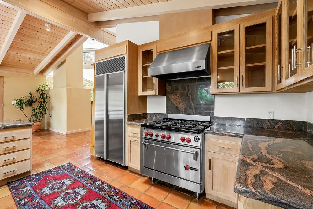 Kitchen with wooden cabinets, stainless steel appliances, dark granite countertops, tiled floor, and a colorful patterned rug.