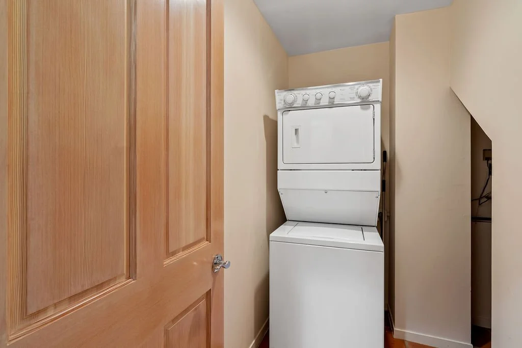 A stacked white washer and dryer in a small laundry closet with beige walls and a wooden door.
