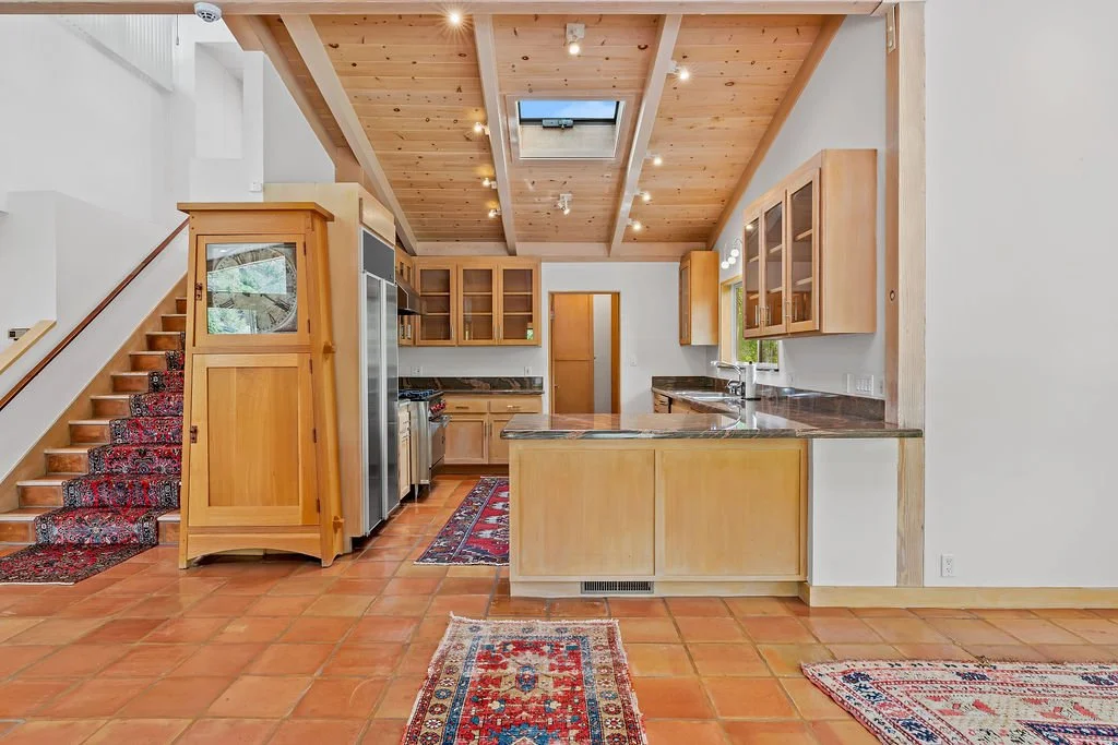 Open kitchen with wooden cabinets, tiled floor, and ceiling with wooden beams and skylight. Stairs with a decorative runner carpet lead up on the left side.
