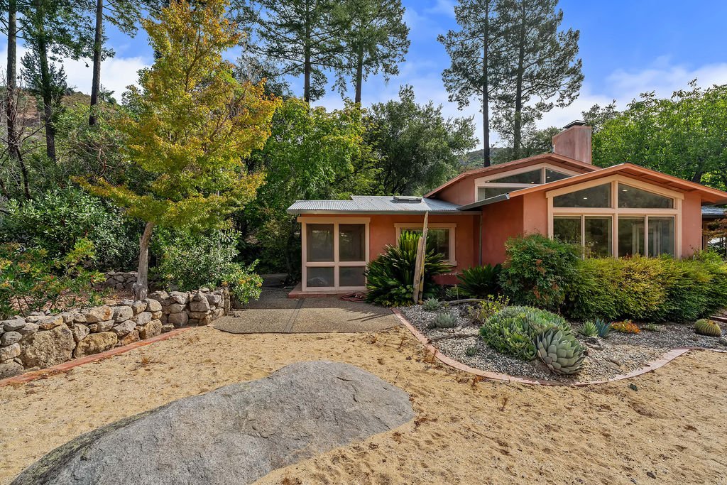A house with large windows surrounded by desert landscaping, including succulents, rocks, and trees.