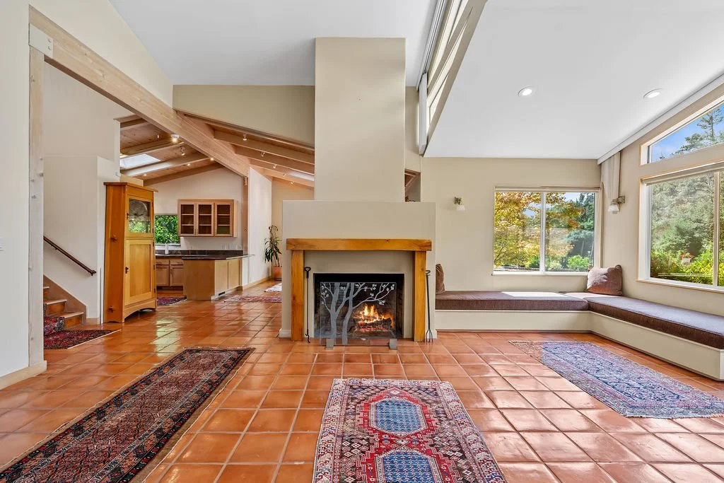 Living room with a fireplace, built-in window seats, large windows showing trees, and a view of the kitchen in the background.