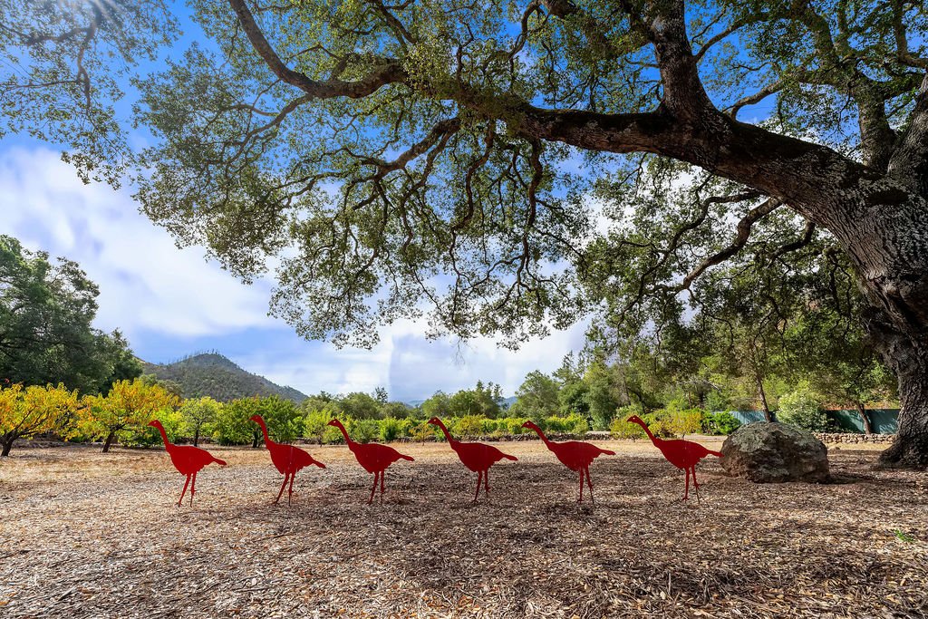 Six red cartoonish silhouettes of dinosaurs with long necks are standing under a large tree in an outdoor setting, with a mountain in the background and a partly cloudy sky overhead.