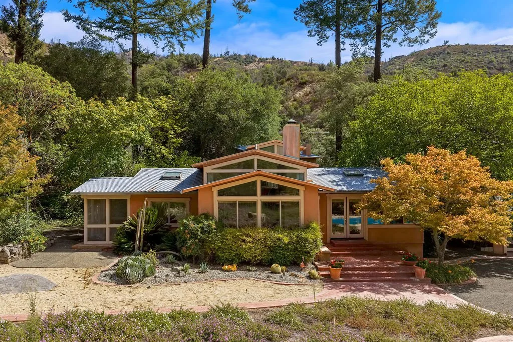 A modern house with large glass windows, orange exterior walls, a metal roof, surrounded by trees and desert plants, with a red brick pathway leading to the entrance.