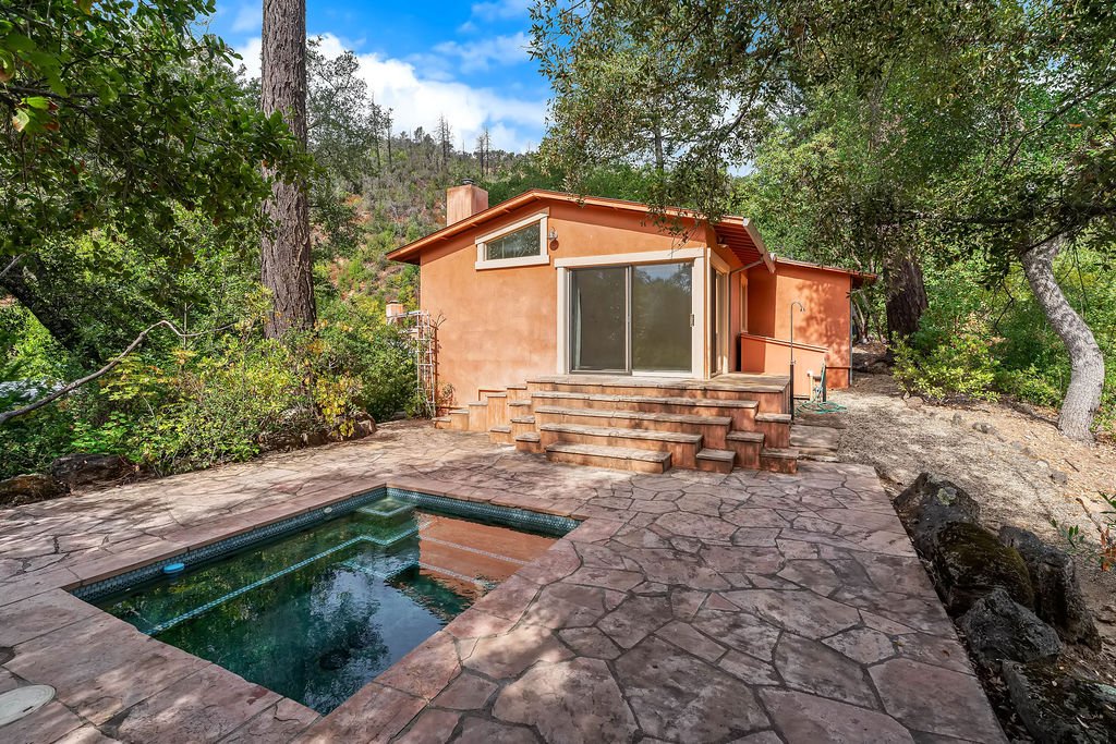 A backyard scene featuring a small house with a stucco exterior, large sliding glass door, and steps leading up to it. There is a nearby hot tub and a stone patio surrounded by trees and greenery.