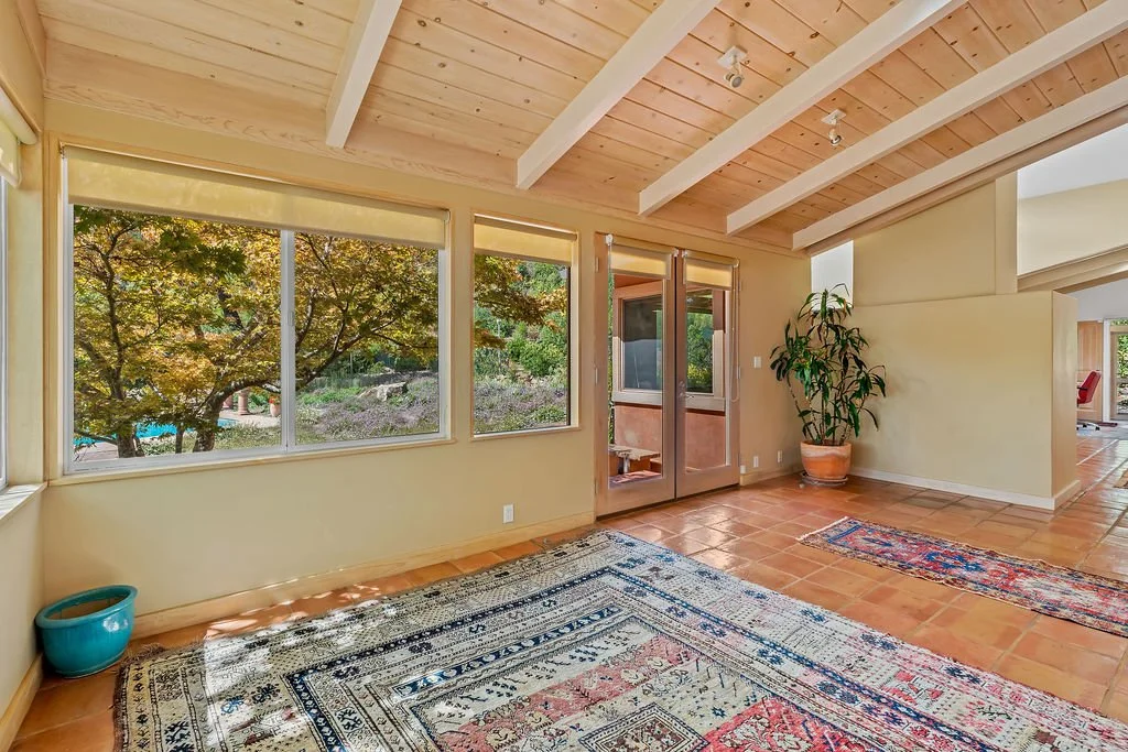 Bright living room with large windows overlooking trees, terracotta tile floor, and a potted plant in the corner.