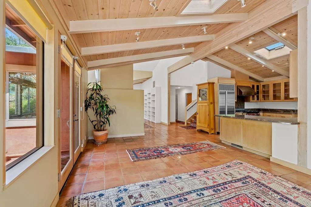 Open-concept living space with wood-paneled vaulted ceiling, large windows, tiled floor, and a kitchen with wooden cabinets and stainless steel appliances.
