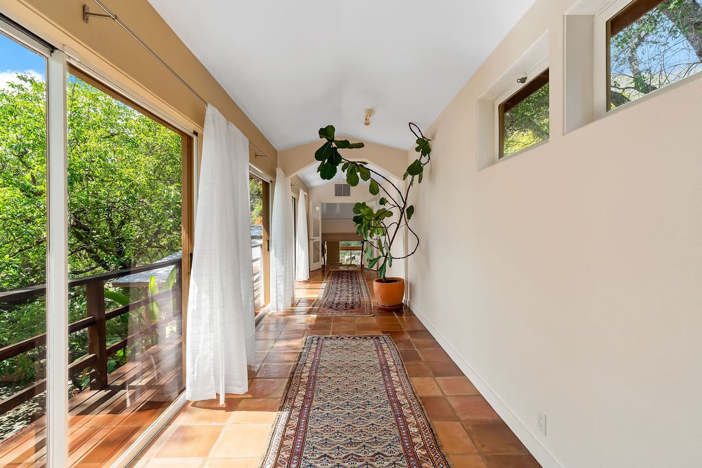 A bright hallway with large sliding glass doors on the left opening to a balcony with green trees outside. White curtains hang beside the doors. On the right wall, there are three small rectangular windows. The floor is terracotta tiles covered parti