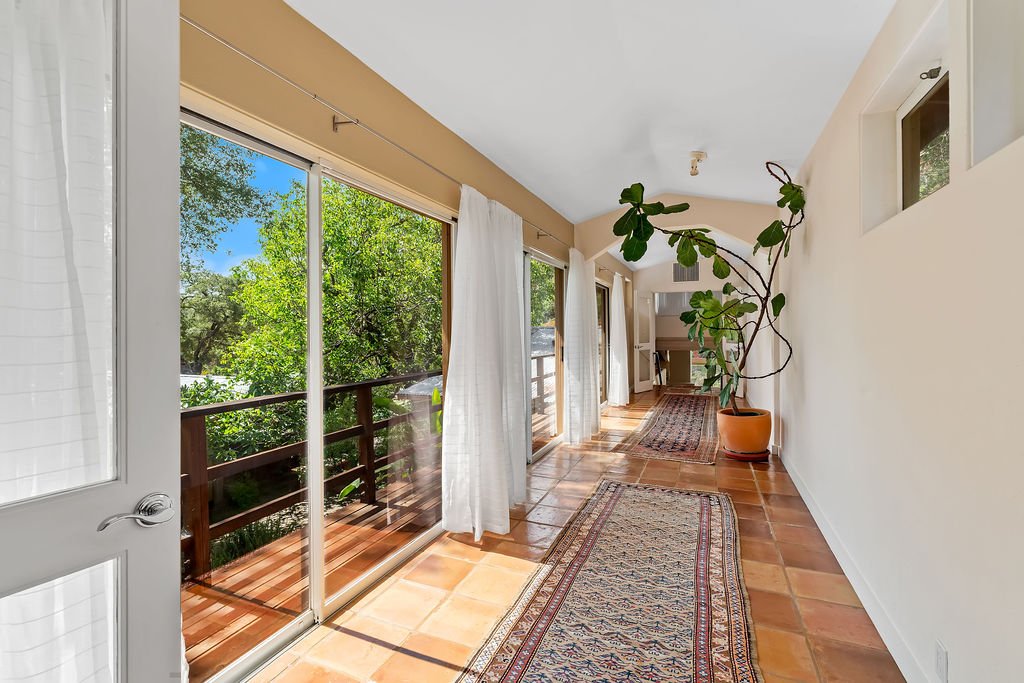 Sunny room with large sliding glass doors leading to a balcony, with white curtains, a potted fiddle leaf fig plant, a patterned rug, and wooden tile flooring.
