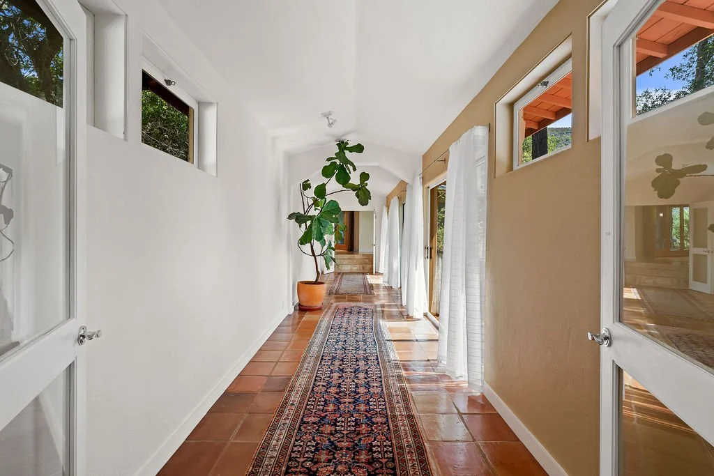 Bright hallway with terracotta tile flooring, white walls, large potted plant, and large windows with curtains allowing natural light.