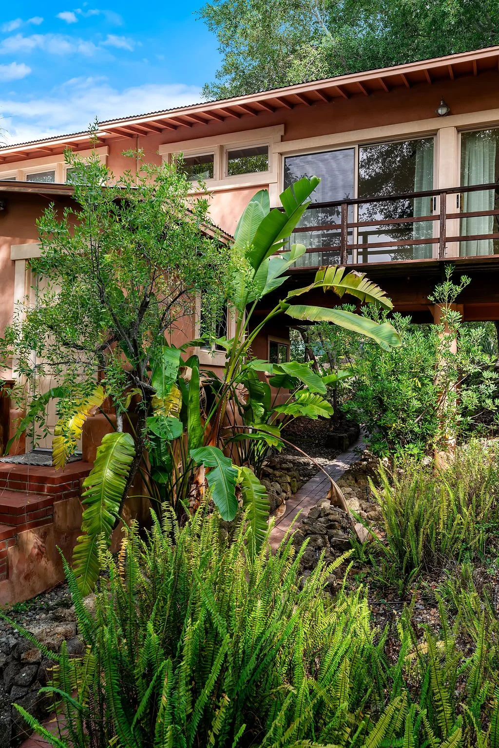 A multi-story house with a balcony and large glass windows, surrounded by lush green tropical plants, including banana plants and ferns, under a partly cloudy sky.