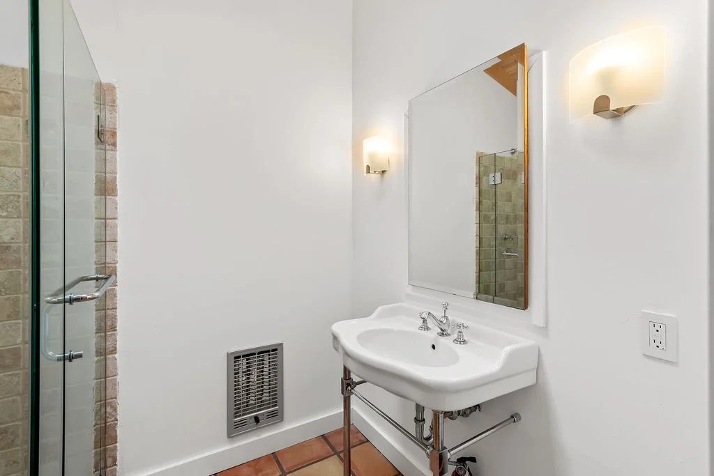 A bathroom with a white wall, a small vintage sink with silver fixtures, a large mirror, two wall-mounted lights, a vent on the wall, a glass shower door with tiled walls visible inside