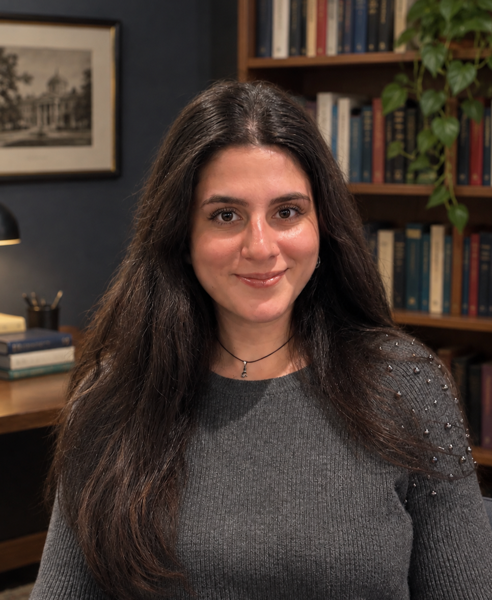 A woman with long dark hair and a gray sweater with embellishments on the shoulder, smiling in a room with bookshelves and framed art.