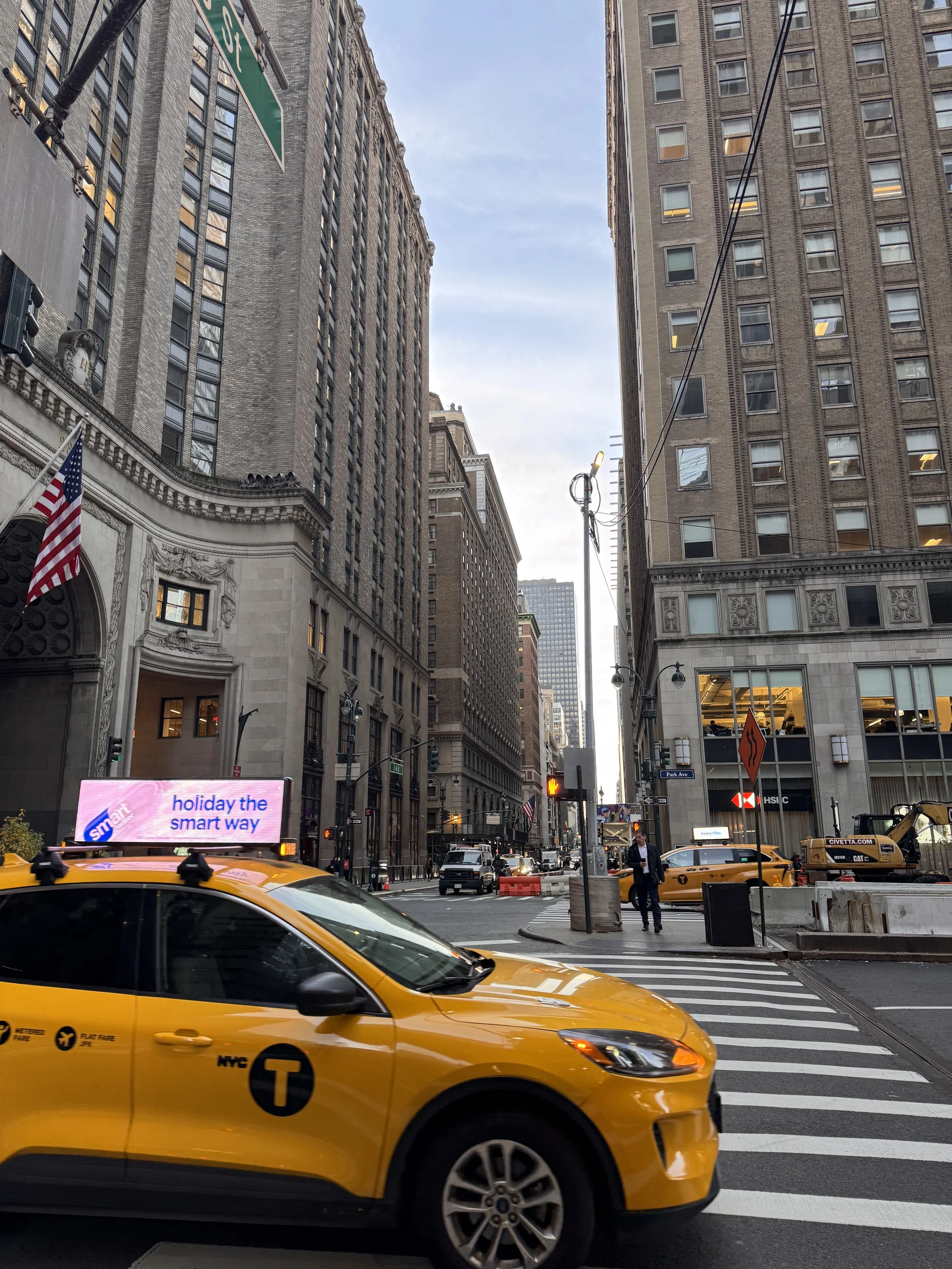 City street with tall buildings, yellow taxis, a pedestrian, an electronic billboard, and street signs. The sky is partly cloudy.