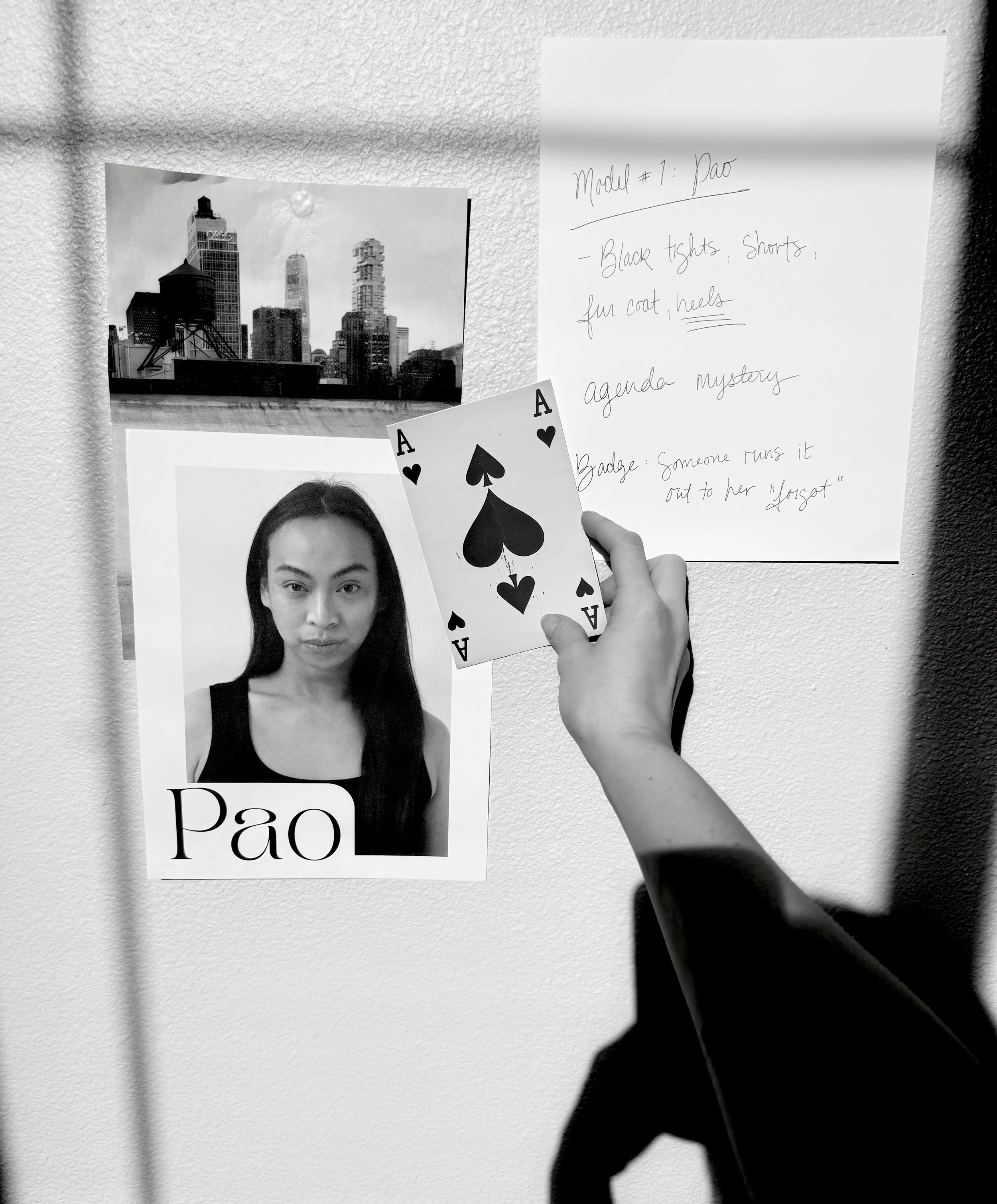 A black and white image of a vision board featuring a fashion model, a magazine clipping of a city, and some notes taken by hand. There is an arm holding up an ACE playing card. This is for Muse Behavior Runway.