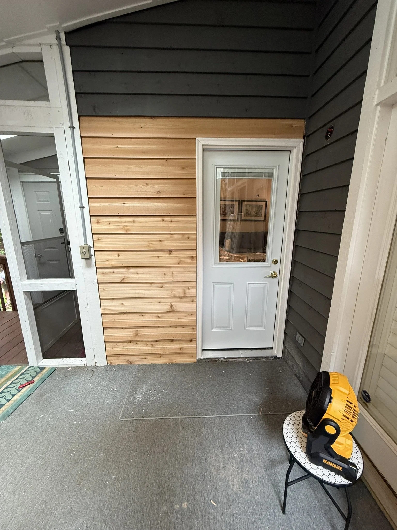 Indoor porch or deck area with a door, wood siding, and construction tools including a yellow DeWalt nail gun on a small round table with a hexagon pattern. The floor is carpeted, and there is a mirror or glass panel on the door.