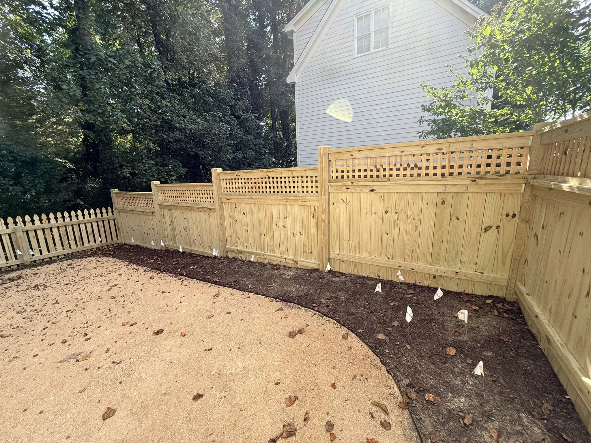 A wooden fence surrounds a backyard area with a light-colored house and deciduous trees in the background. The ground in the yard is partially covered in dirt and fallen leaves, with some white markers along the perimeter of the fenced area.