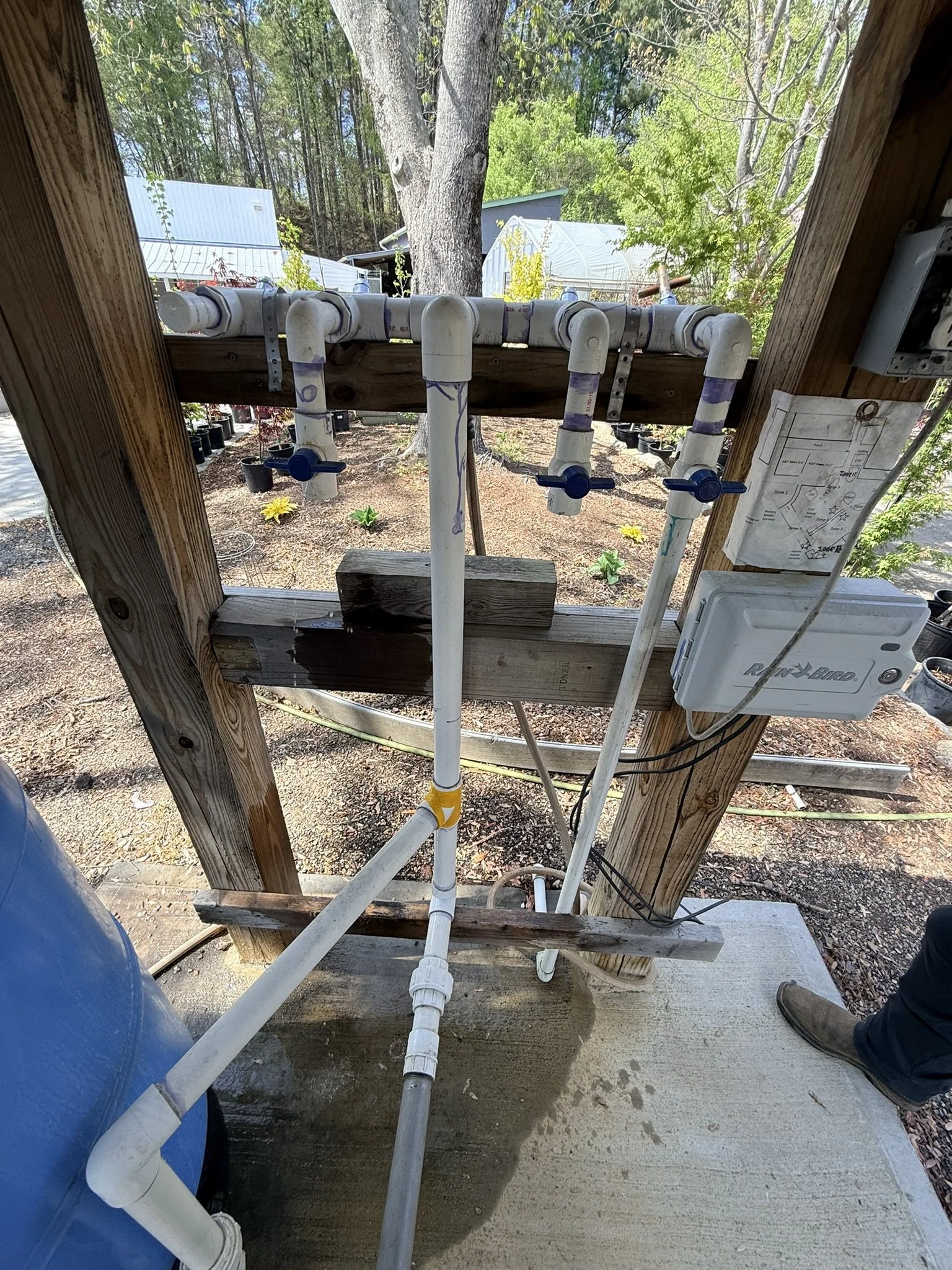 A view of plumbing pipes and valves installed under a wooden structure, with a tree and outdoor landscape in the background.