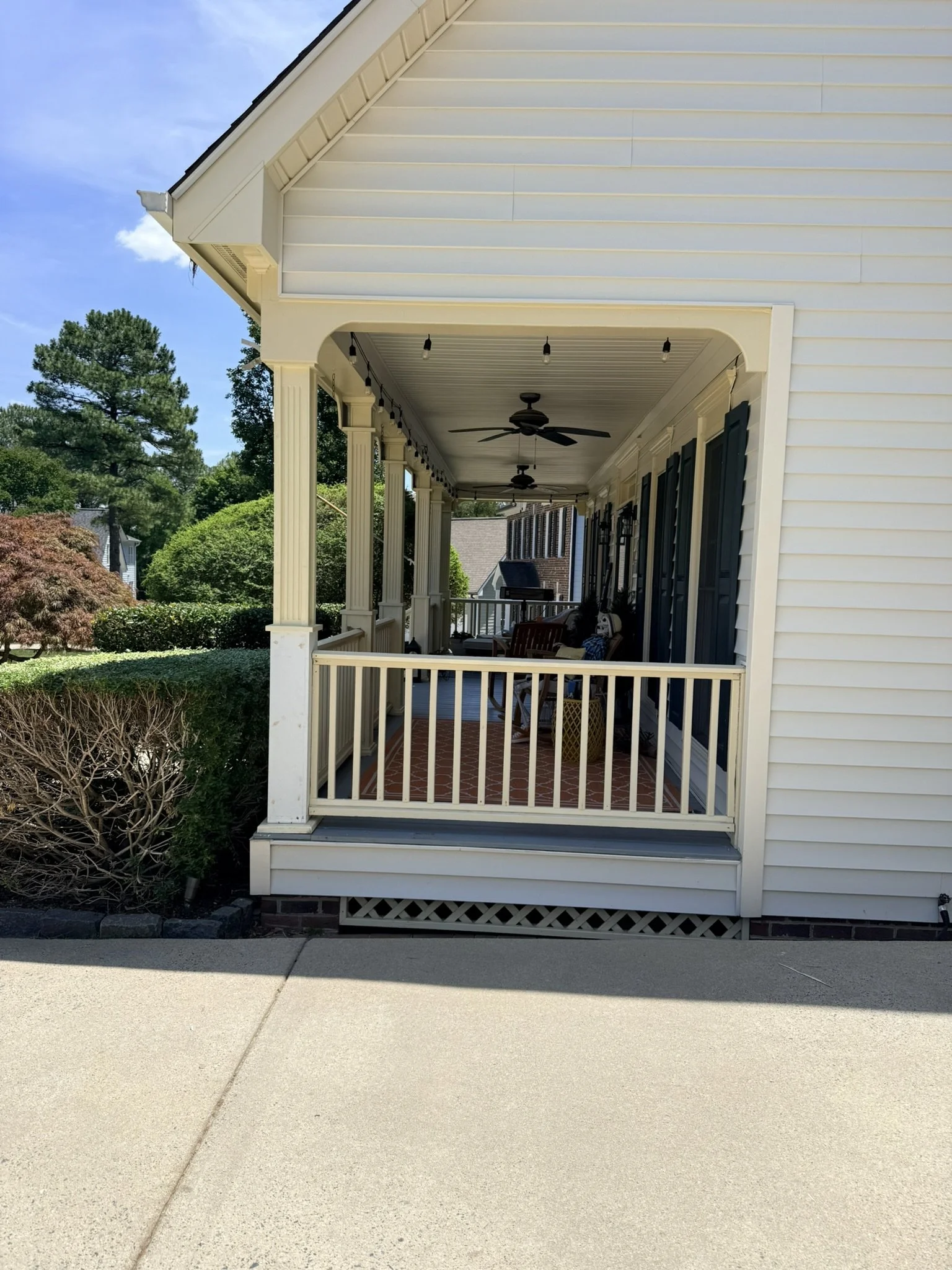 A view of a white house porch with black shutters, ceiling fans, and string lights, surrounded by greenery and trees, with a clear blue sky in the background.