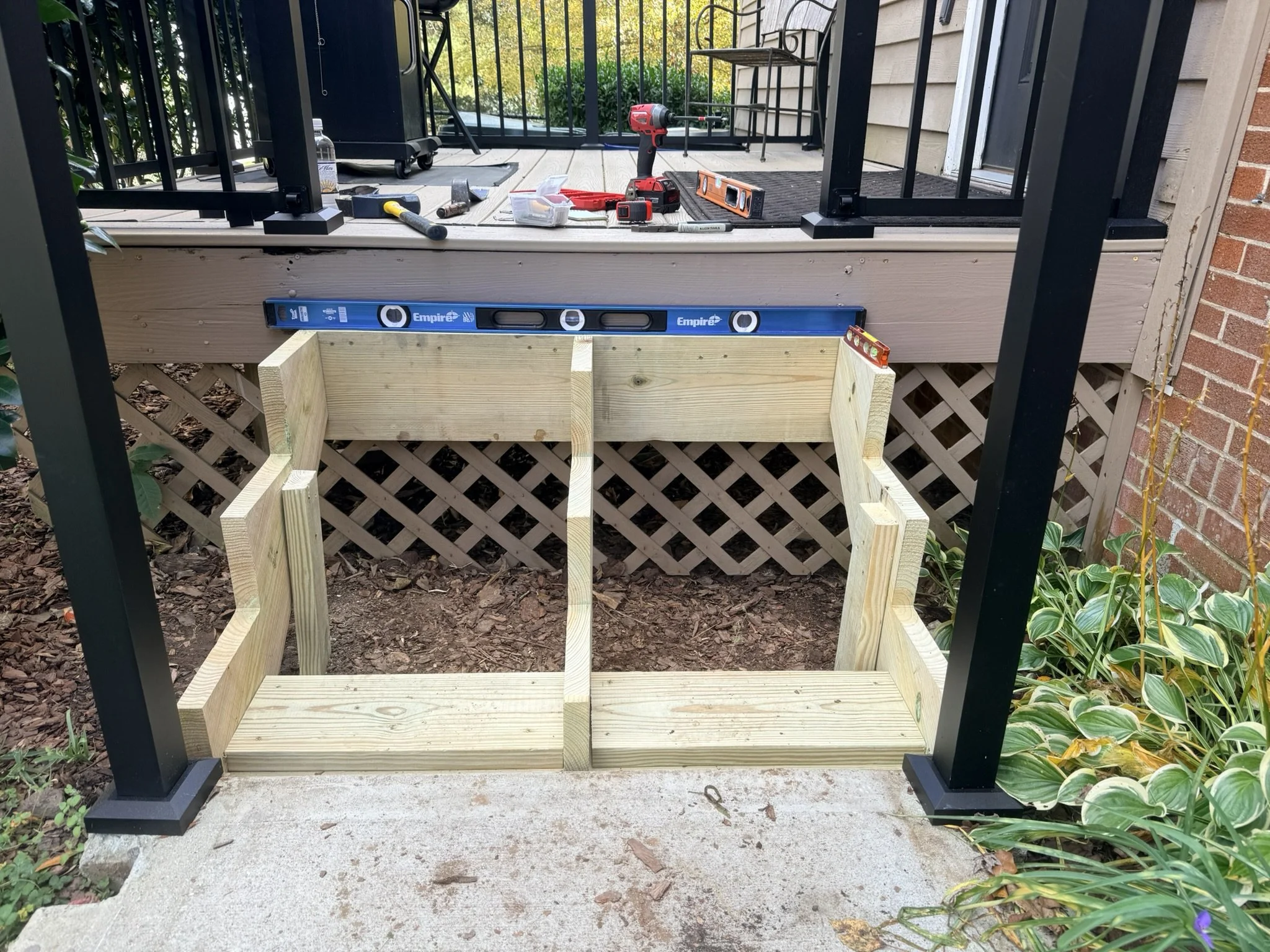 Construction of new deck stairs with black metal railing, tools including level, drill, hammer, and leveler, on the deck surface, with a brick wall and greenery in the background.