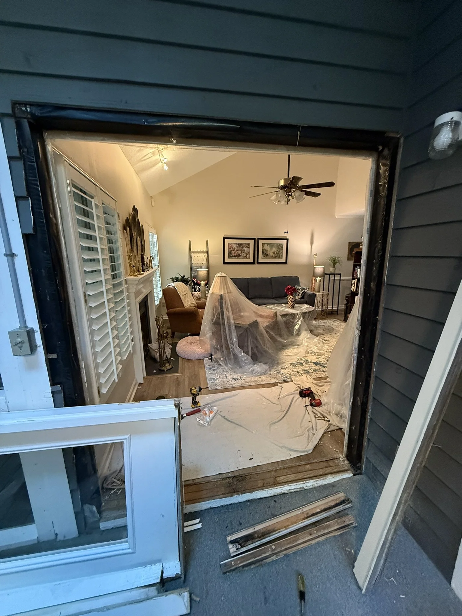 View from outside a house showing a partially removed door and window frame, construction tools, and debris on the ground.