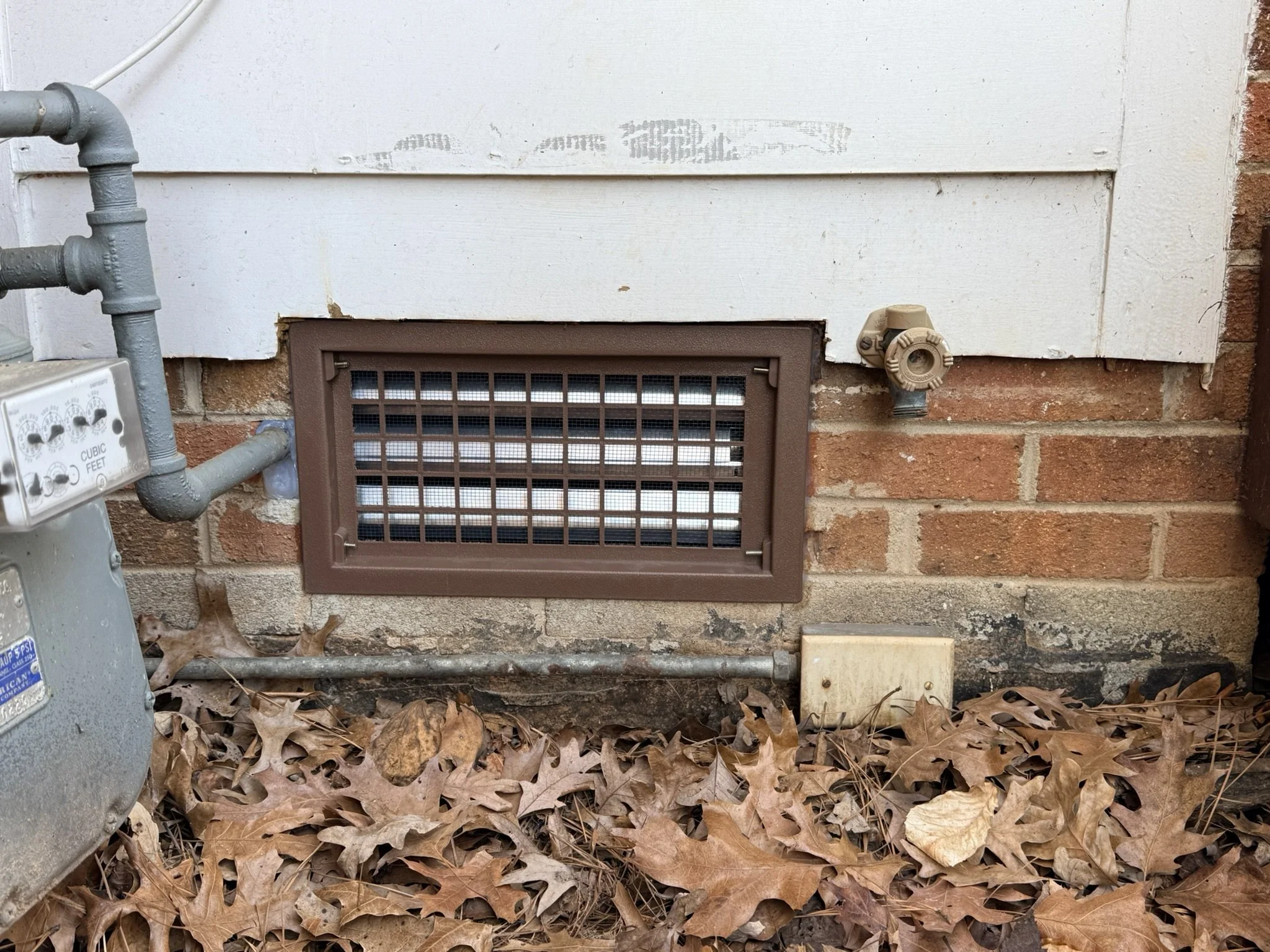 Side of a brick house with an unfinished vent, an outdoor electrical box, and pipes, surrounded by fallen brown leaves.