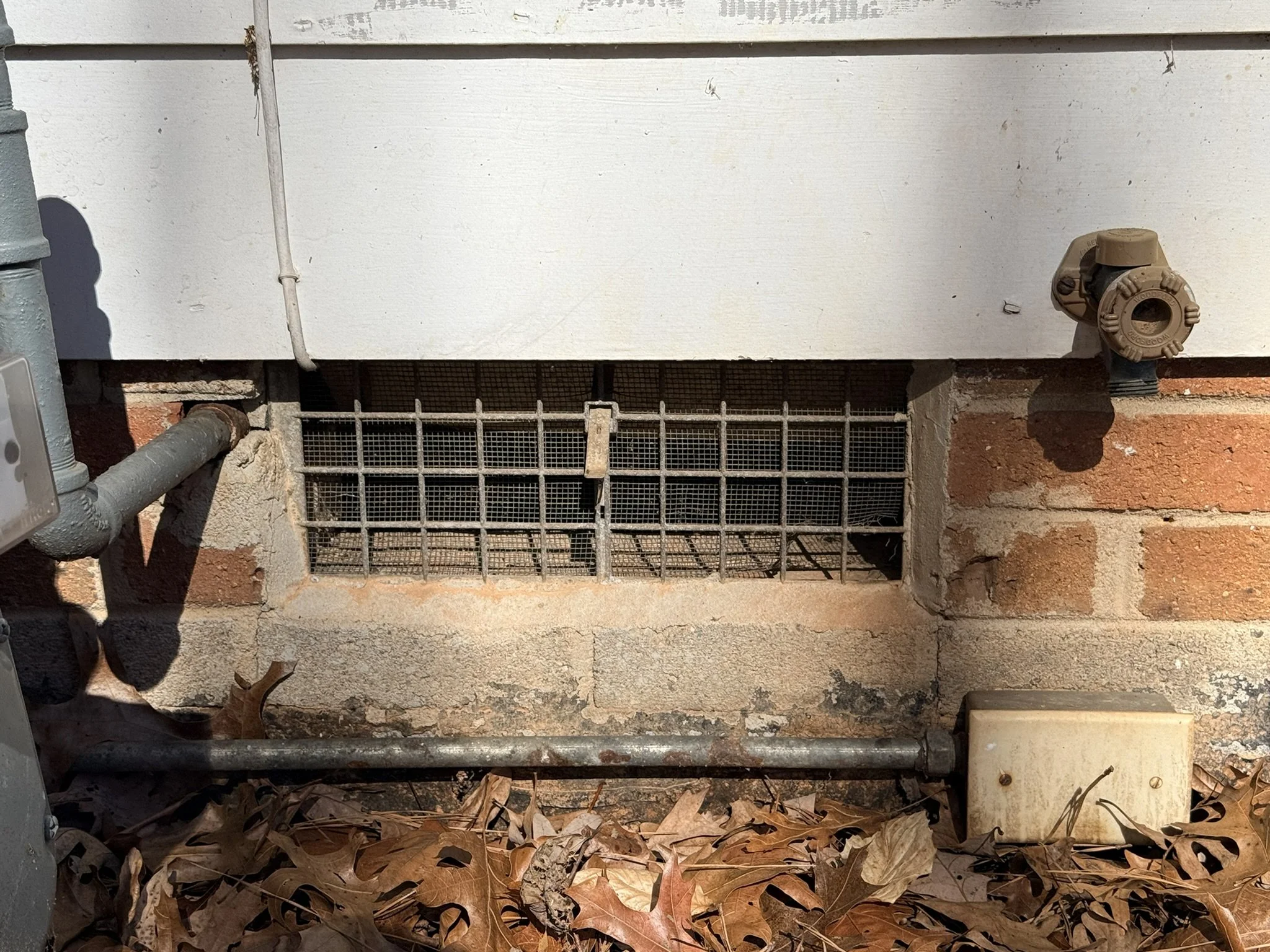 The image shows the bottom part of a building's exterior wall, with a white-painted surface, a metal grate covering an opening, and various pipes and a water valve attached to the wall. There are fallen leaves on the ground in front of the wall.