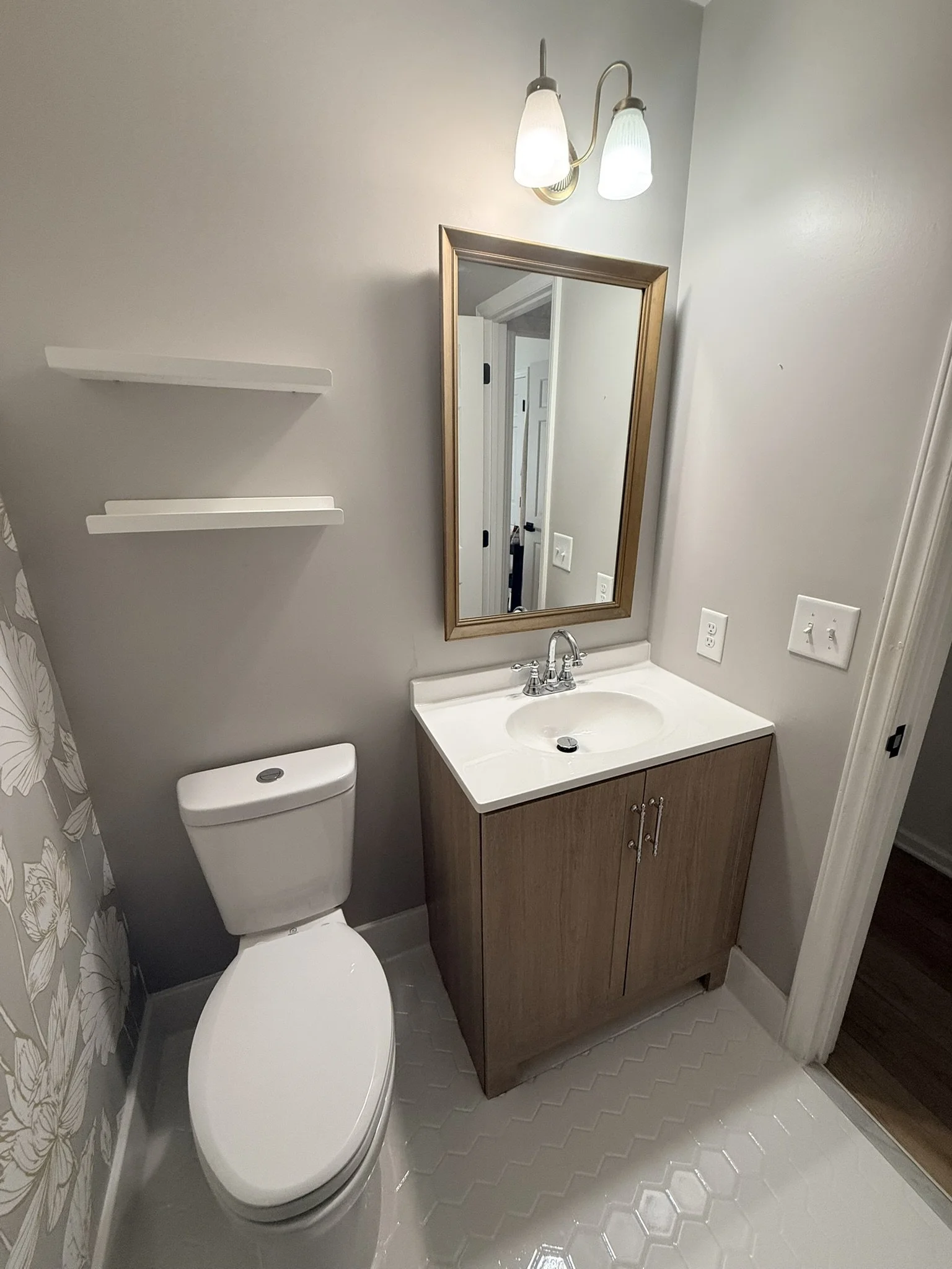 Small bathroom with a white toilet, a cabinet with a sink, a mirror, and a light fixture above the mirror. Two white shelves are on the wall to the left of the mirror. The wall is painted a light gray.