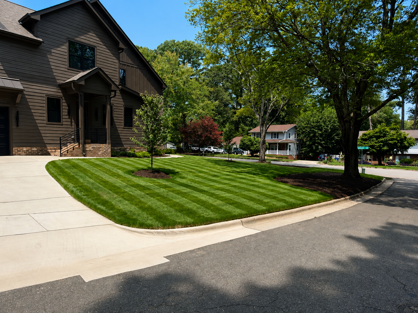 A residential neighborhood with a well-manicured lawn, young trees, and mature trees, along with houses and parked cars in the background.