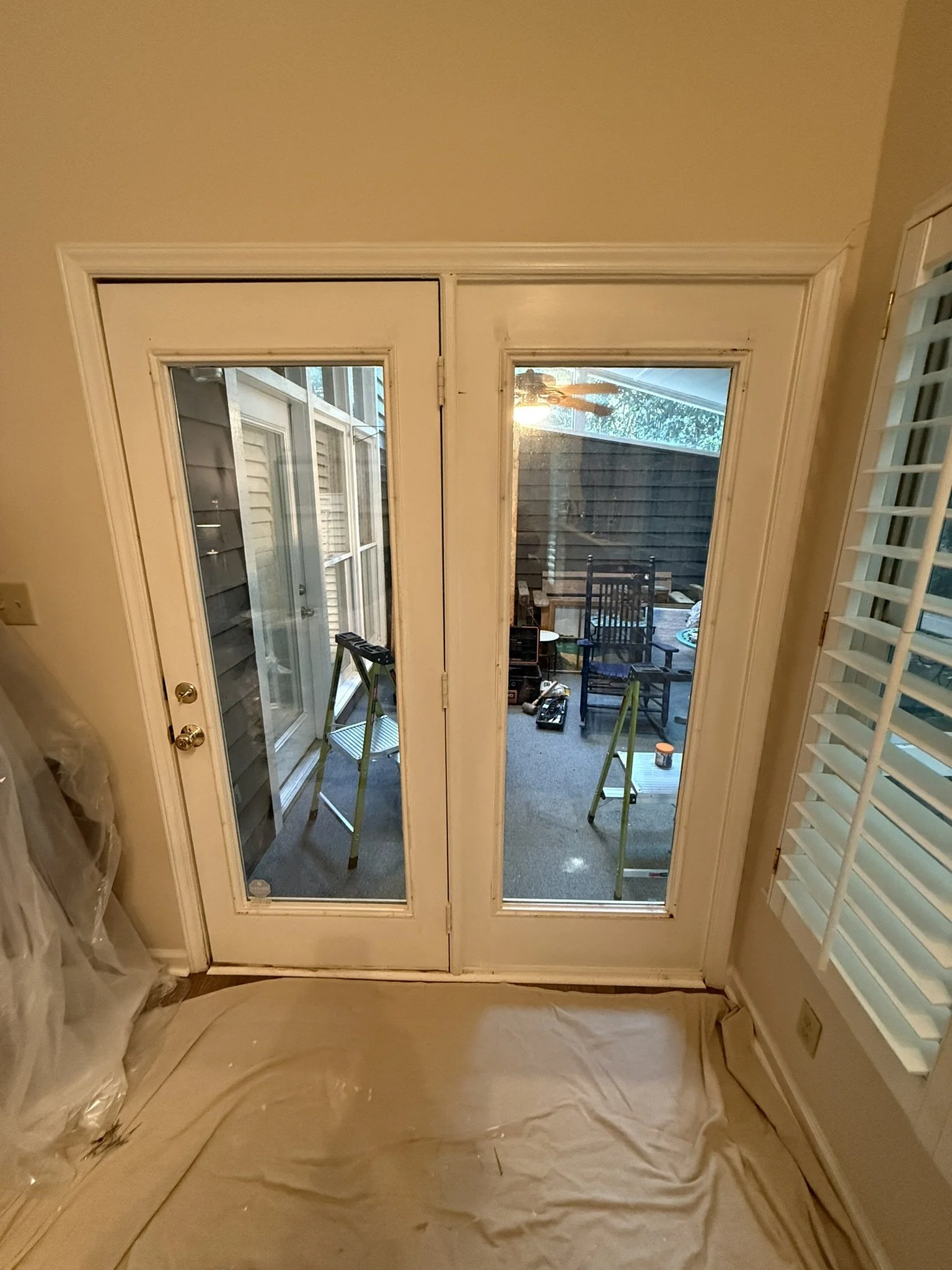 French doors leading to an outdoor patio with chairs, tools, and supplies for renovation, with window shutters on the right.