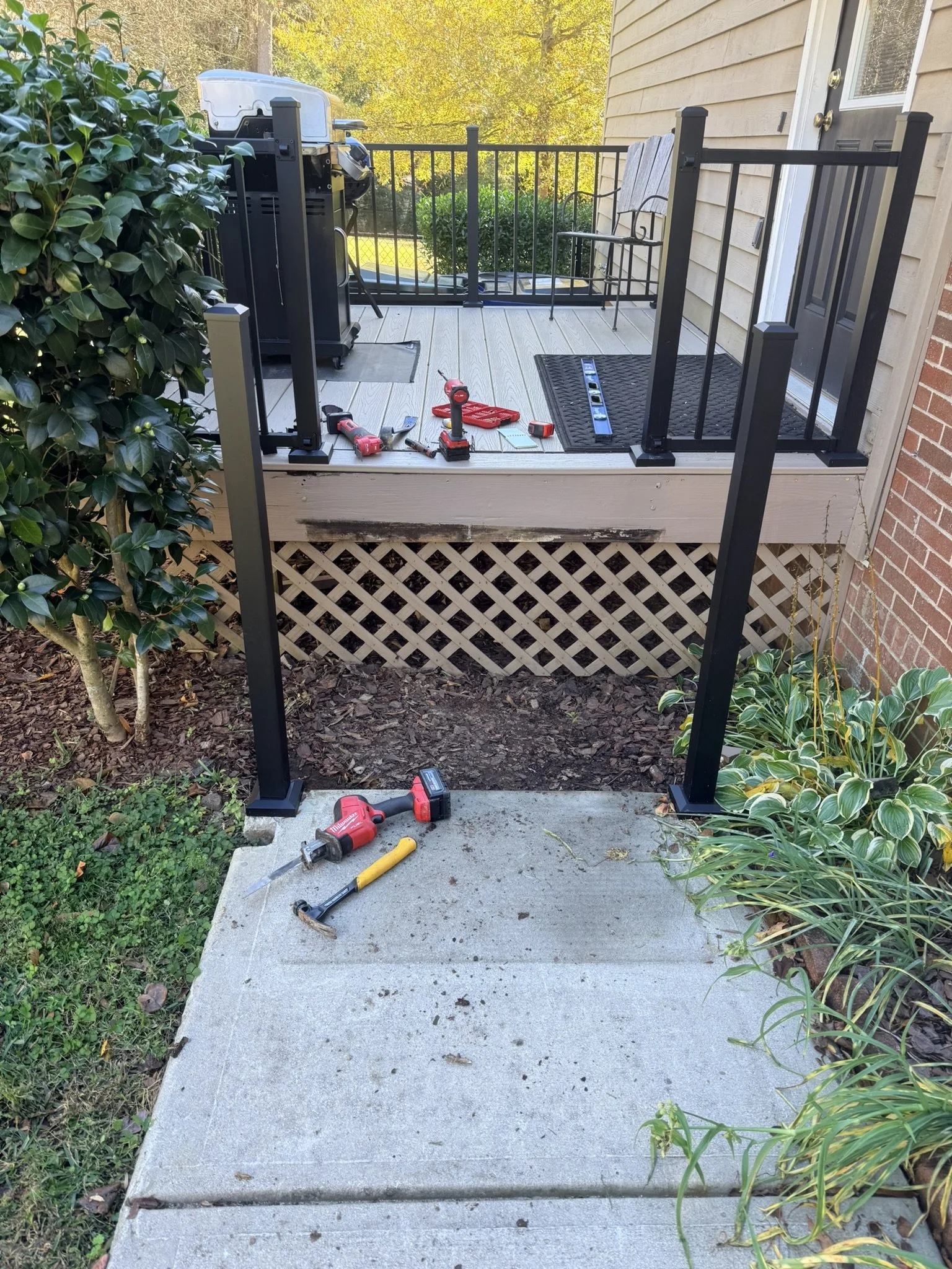 A partially assembled black metal railing on a wooden porch attached to a house, with construction tools such as a cordless drill, level, hammer, and measuring tape scattered on the concrete steps and porch floor, surrounded by bushes and plants.