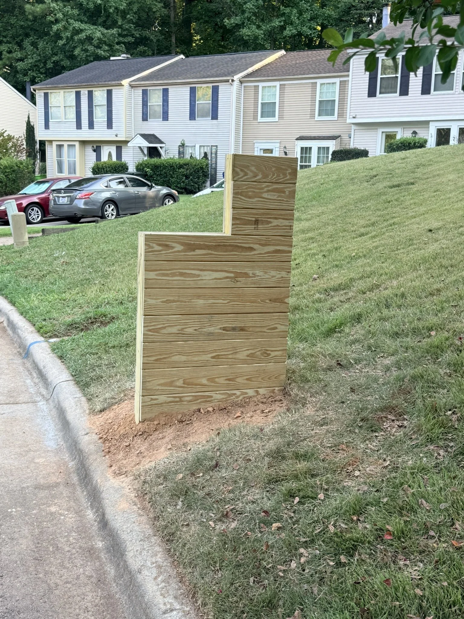 A wooden mailbox is installed in a grassy area near a sidewalk, with residential townhouses and parked cars in the background.