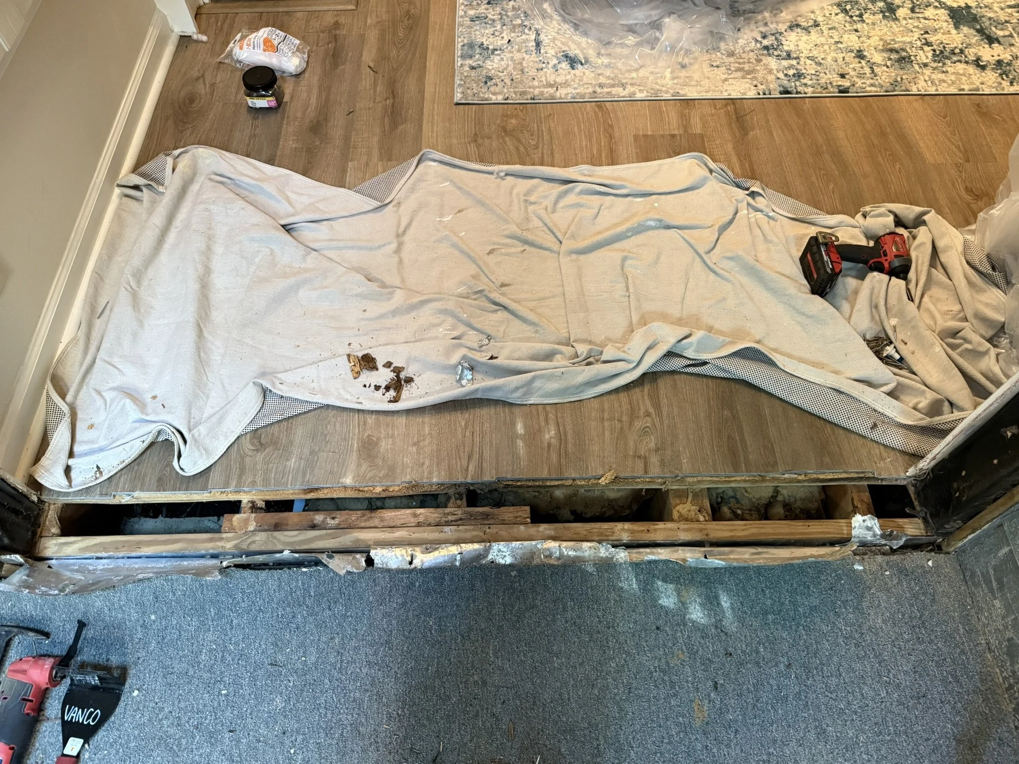 Disassembled wall and flooring with a drop cloth covering the top, a power drill, and a few tools and supplies on the floor, indicating construction or renovation work in progress.