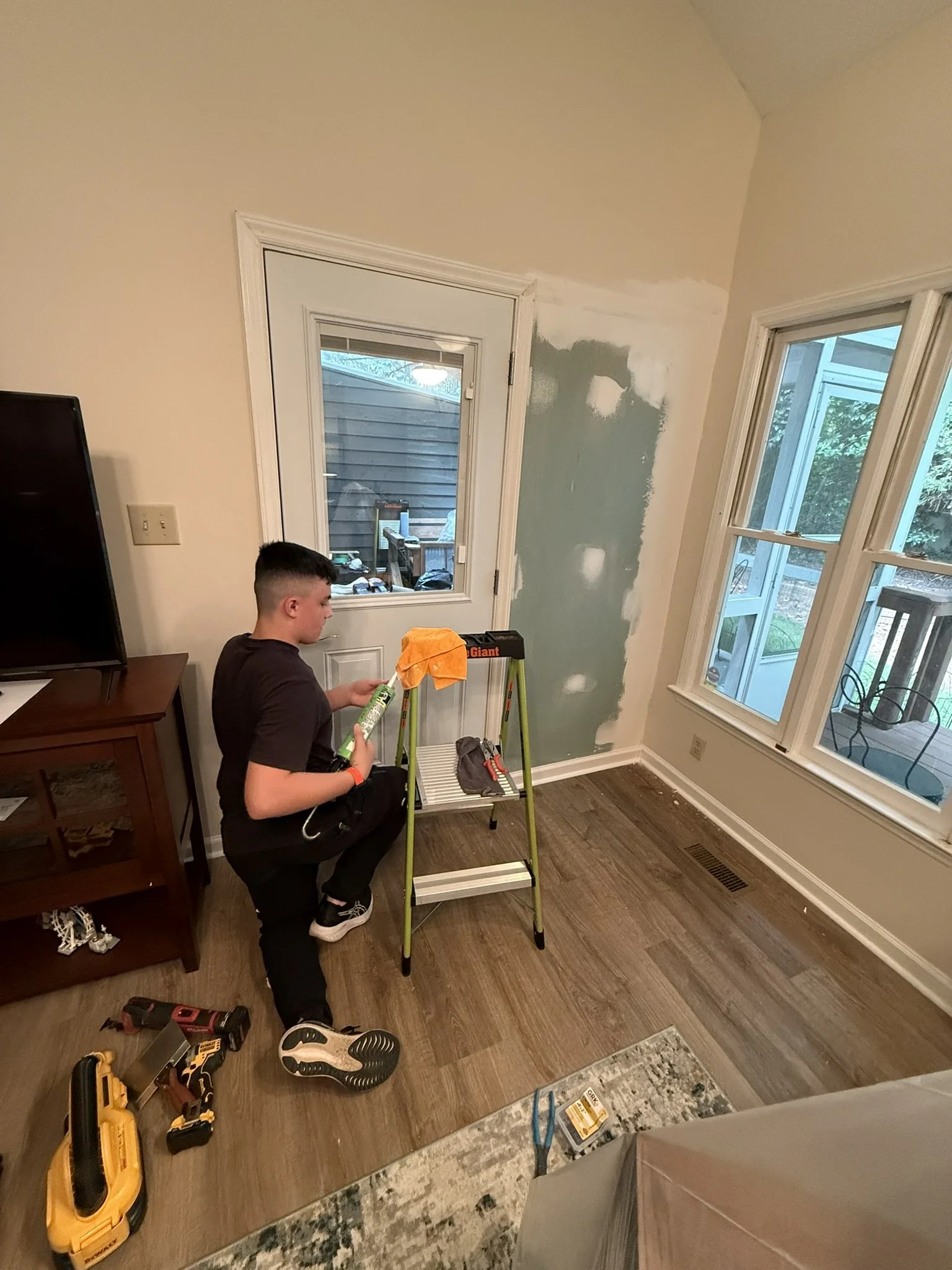 A person kneeling inside a room with light-colored walls and wooden flooring, applying paint to a wall around a window with a roller brush. Tools and supplies are scattered on the floor, including a yellow toolbox, a drill, and a paint tray. The room